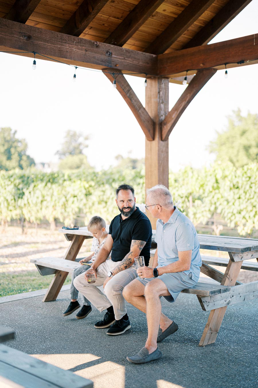Three people sitting on a wooden bench under a gazebo, enjoying drinks and conversation, with a vineyard in the background.