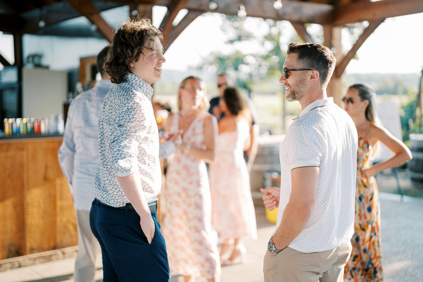 Chatting guests enjoying an outdoor summer party under a wooden gazebo, dressed in casual attire with sunny, vibrant surroundings.
