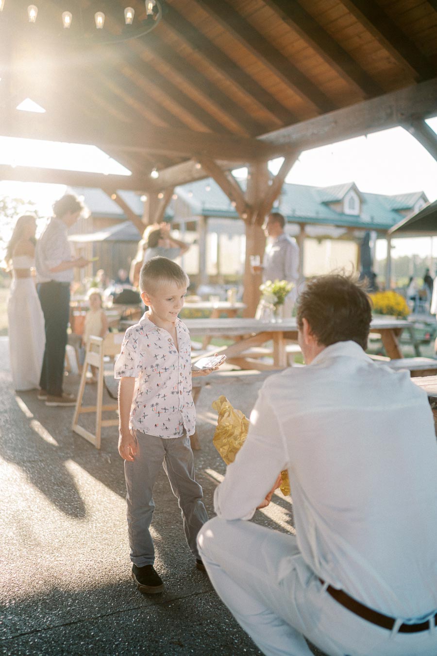 A young boy in a patterned shirt stands under a wooden pavilion interacting with an adult, surrounded by people enjoying an outdoor gathering. Sunlight filters through the structure, creating a warm and inviting atmosphere.