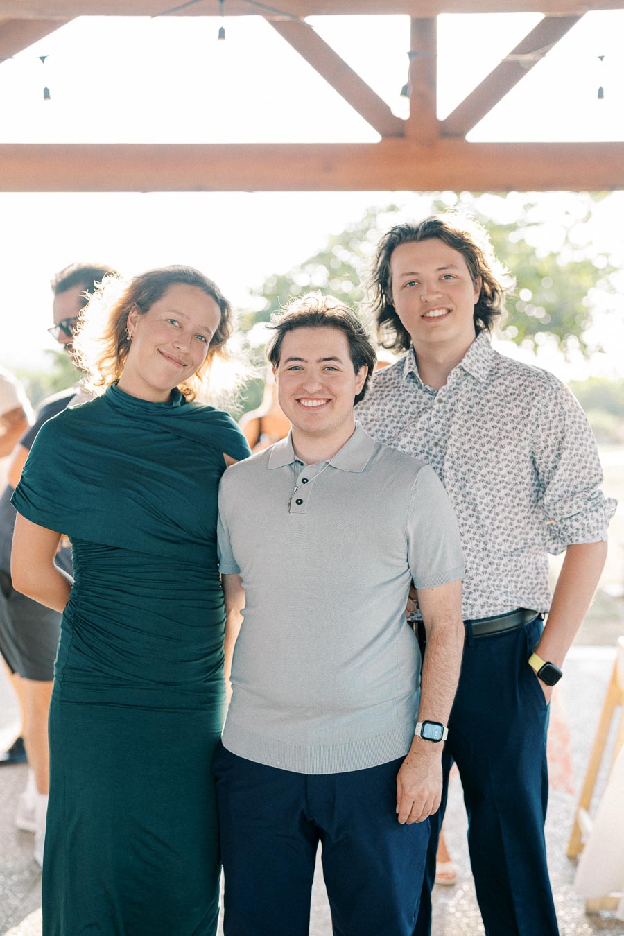 A group of three people smiling outdoors at a social event under a wooden gazebo, with sunlight filtering through and trees in the background. The individuals are wearing casual formal attire, including a green dress and patterned shirts.
