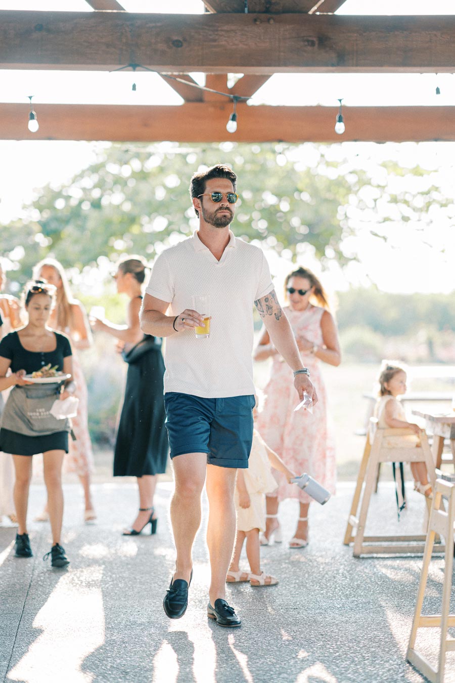 A man in sunglasses and a white shirt holding a drink walks confidently at an outdoor social gathering, surrounded by people mingling in the background under a wooden pergola.