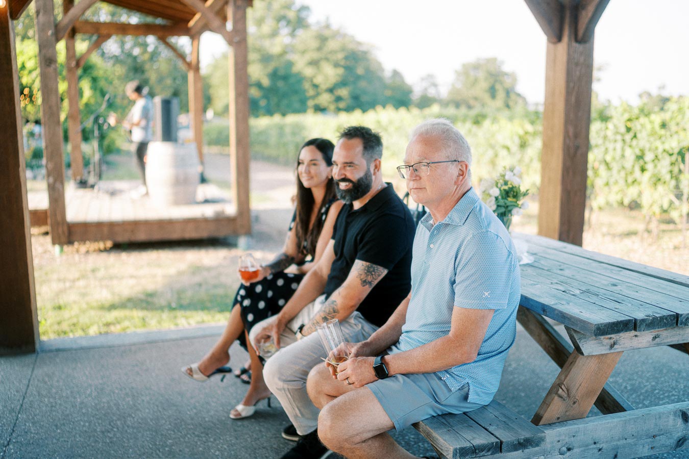 A group of three people sitting on a bench at an outdoor vineyard, enjoying drinks under a wooden pergola. They are facing a musician playing a saxophone in the background, with lush grapevines visible in the distance.