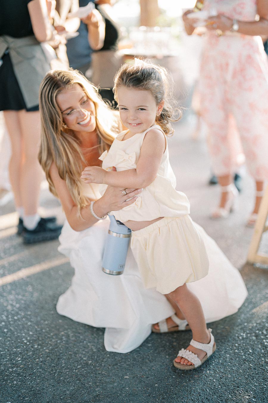 A woman crouching with a smiling young girl holding a drink container in a sunny outdoor setting with blurred people in the background.