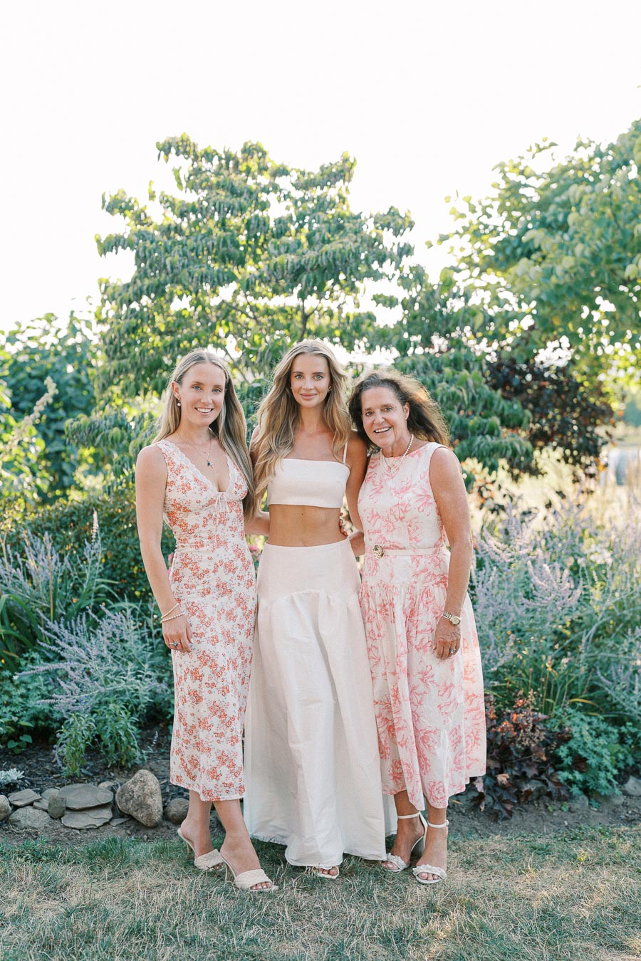 Three women wearing floral dresses and a white crop top pose outdoors in a garden setting.