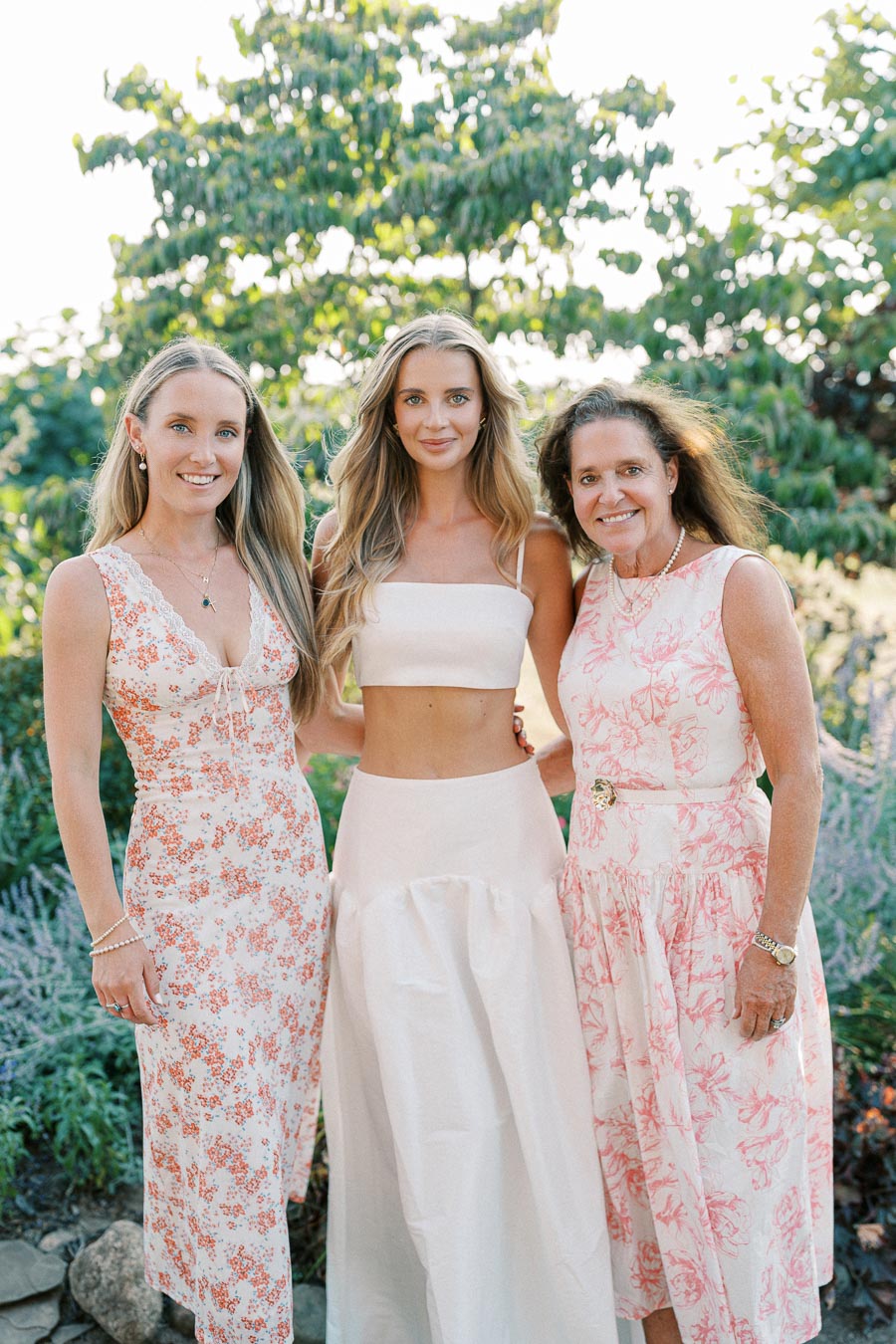 Three women in floral dresses stand together outdoors, smiling against a backdrop of lush green trees.