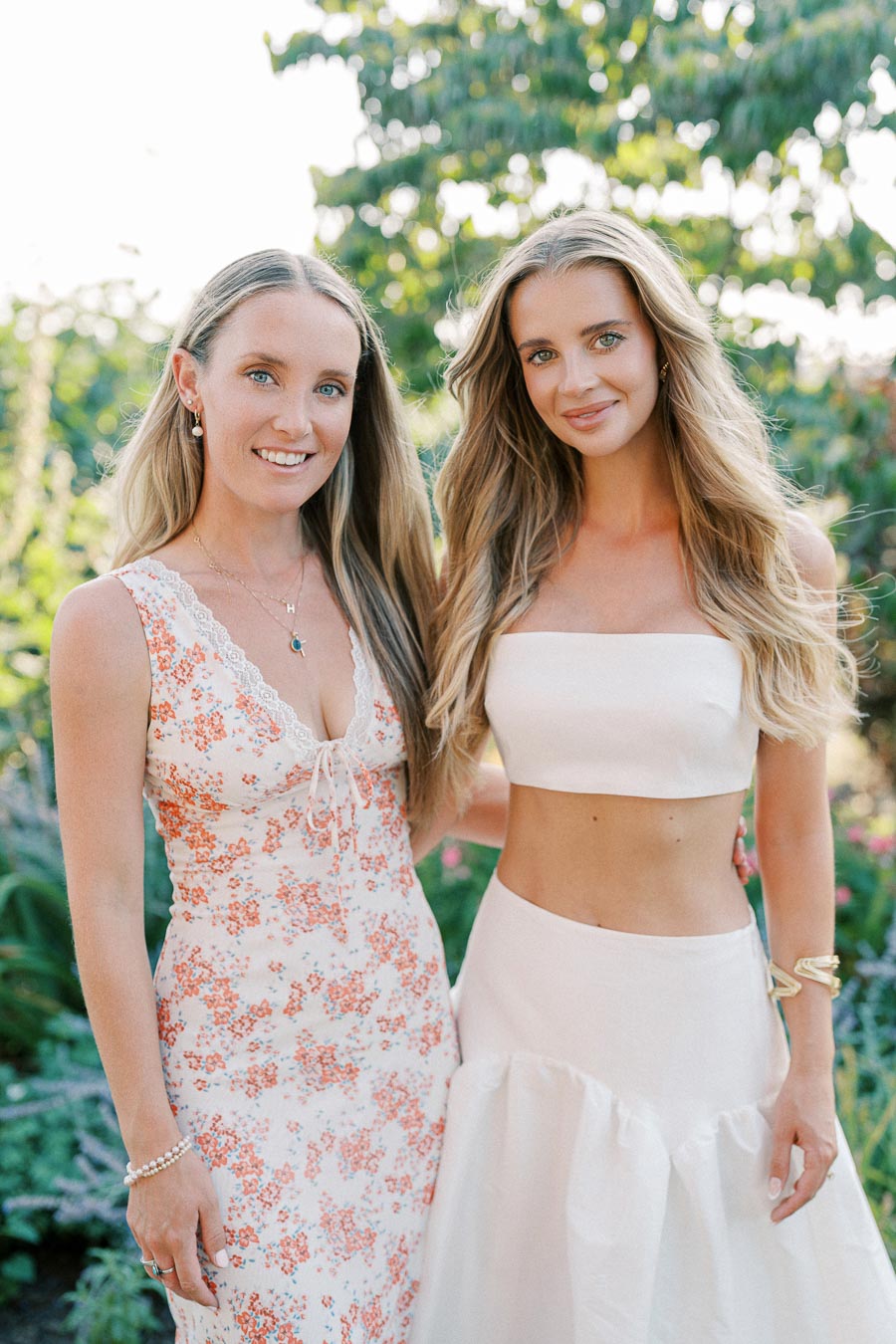 Two women smiling in a garden setting, wearing summer dresses with floral and white patterns, surrounded by greenery and sunlight.