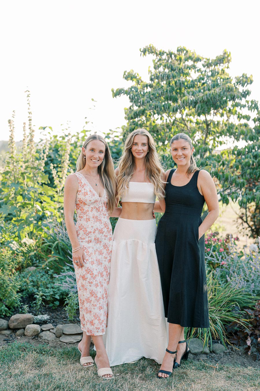 Three women posing in a garden setting, wearing summer dresses. The vibrant greenery and floral background enhance the relaxed, outdoor atmosphere.