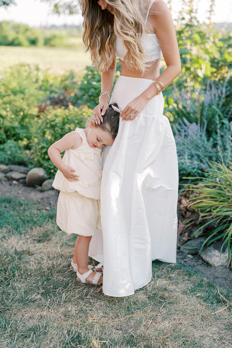 A mother and young daughter in matching white dresses, embracing in a sunny garden setting, showcasing family bonding and summer fashion.