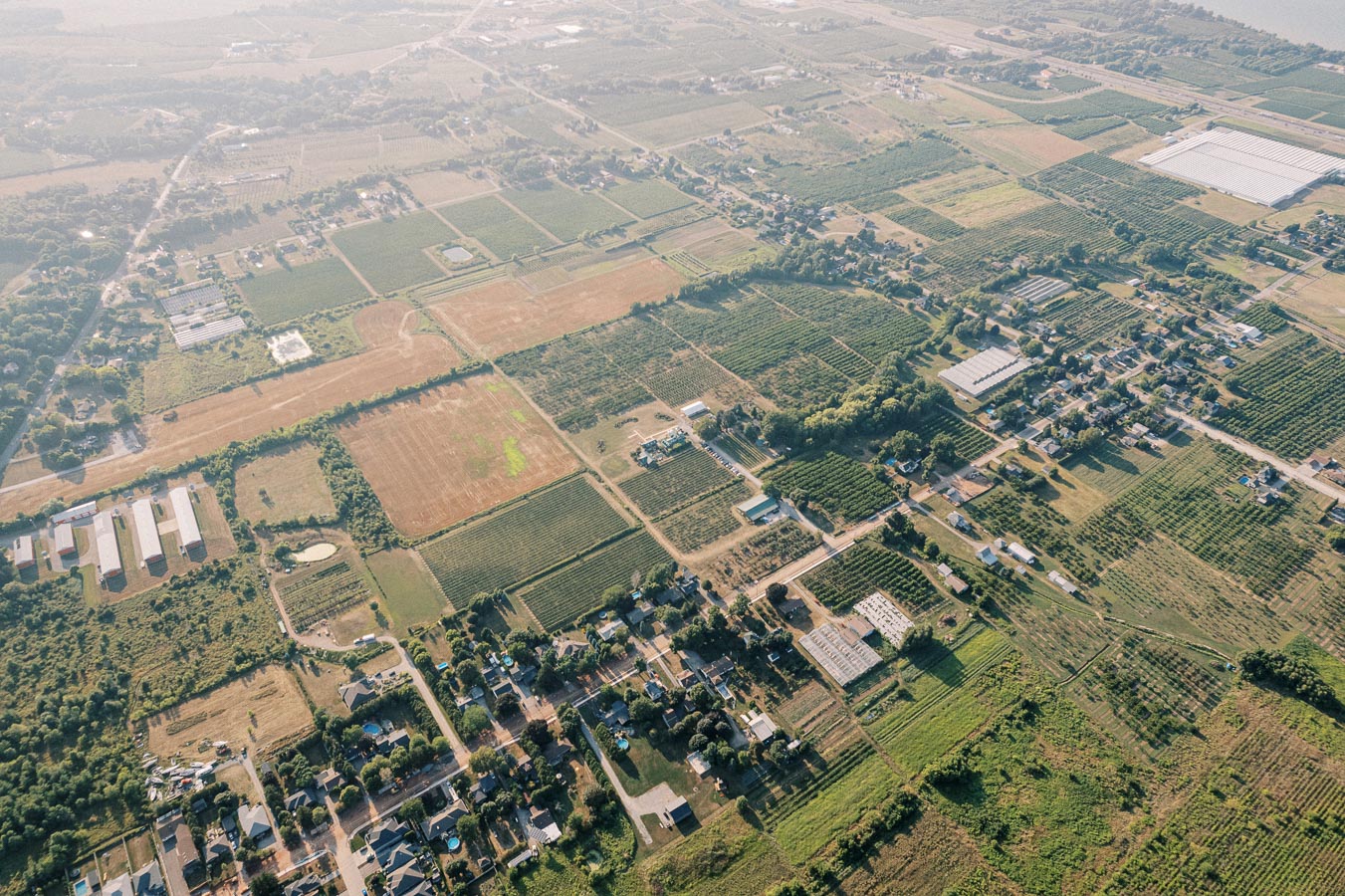 Aerial view of a rural landscape with patchwork fields, residential areas, and greenhouses on a sunny day.