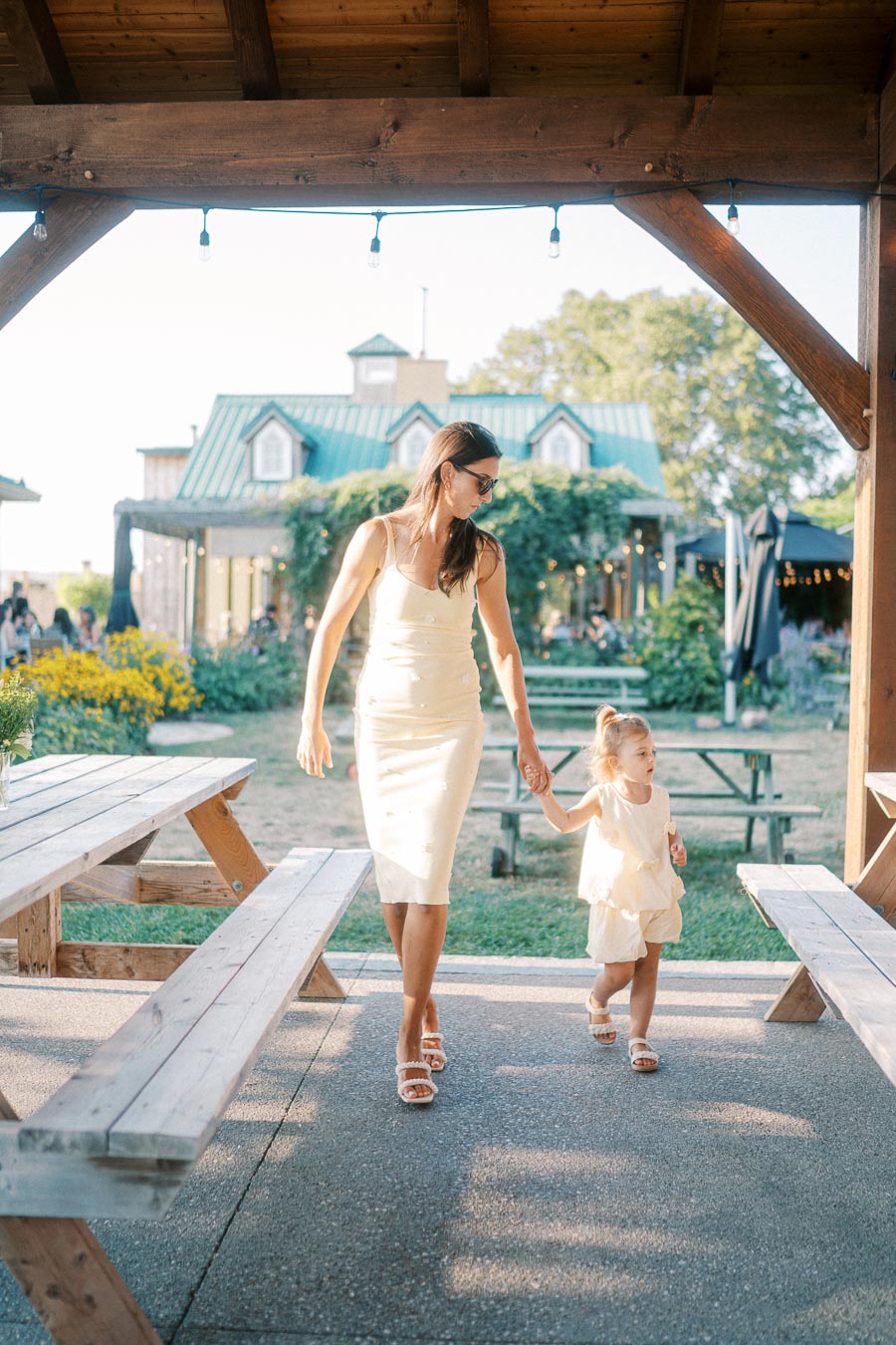 A mother and daughter holding hands walk through an outdoor garden space with wooden picnic tables and a cottage-style building in the background, captured under a wooden pergola on a sunny day.
