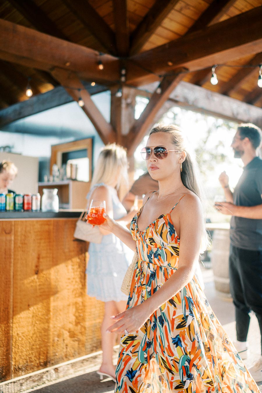A woman in a colorful summer dress holding a drink at an outdoor bar, with people socializing in the background under a wooden roof.