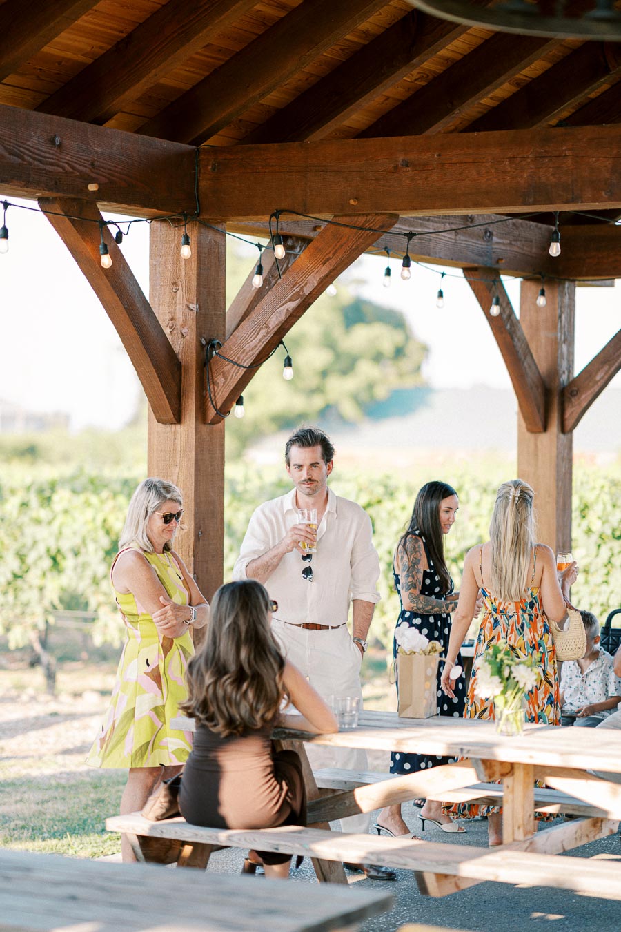 Group of people socializing under a rustic wooden pergola at an outdoor event, enjoying drinks and conversation on a sunny day.
