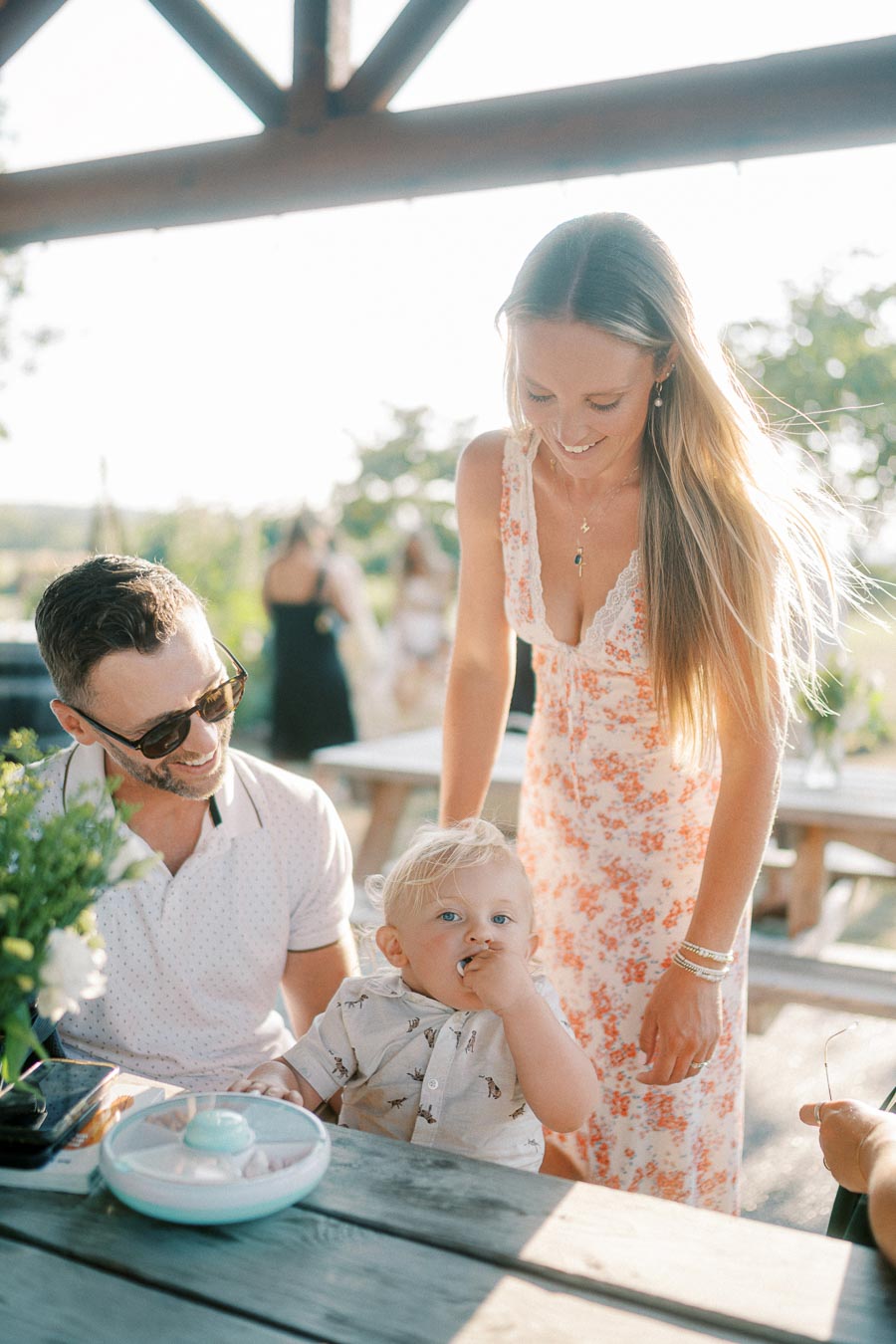 Happy family spending quality time outdoors at a picnic table, with a smiling father in sunglasses and a mother in a floral dress interacting with their young child, who is curiously looking up.