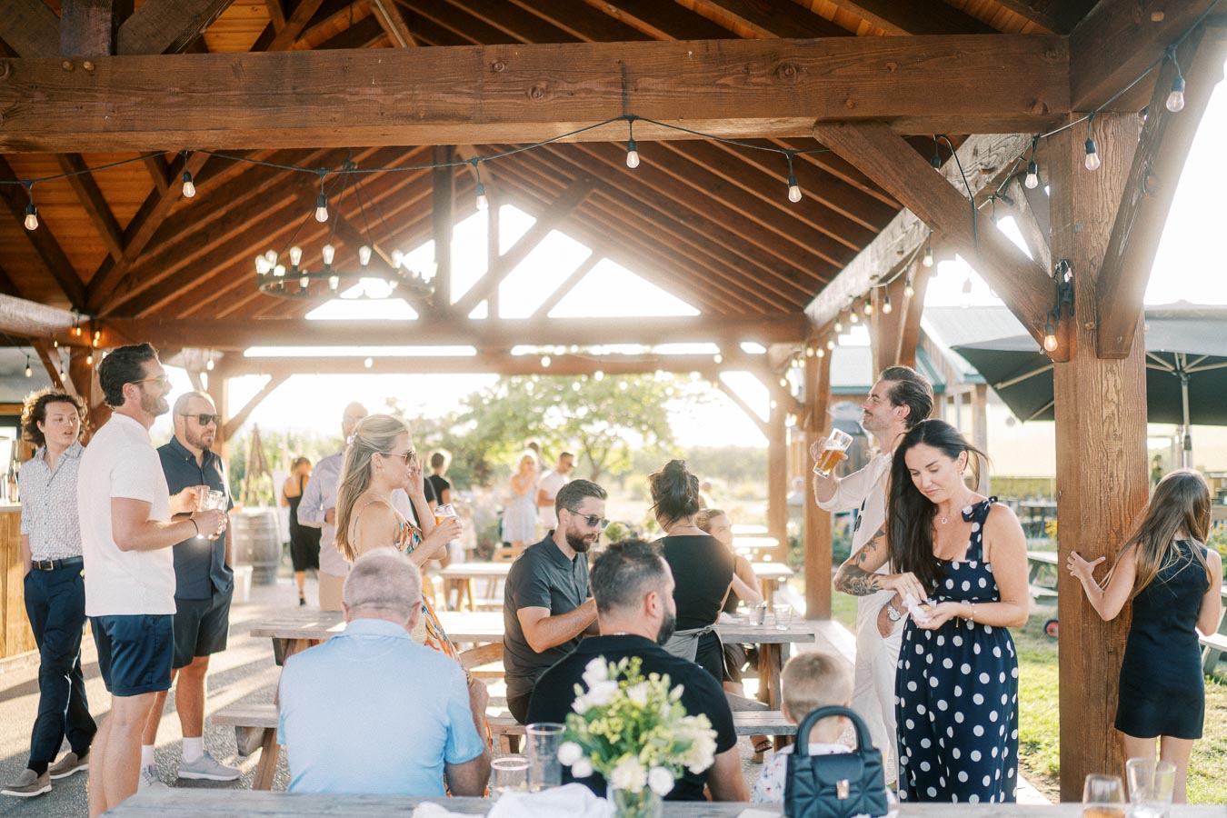 A lively outdoor gathering under a wooden pavilion with string lights, featuring people socializing and enjoying drinks on a sunny day, surrounded by greenery.