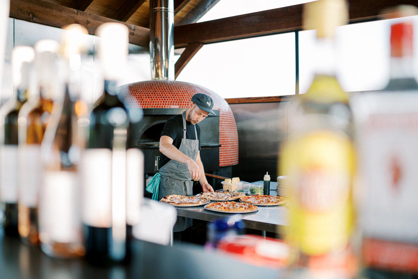 Chef preparing pizzas in a rustic kitchen with a wood-fired oven in the background, surrounded by bottles on a counter.