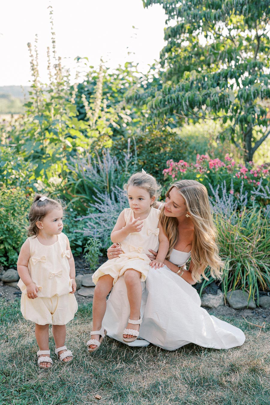 A woman crouches in a lush garden with two young children in yellow dresses, surrounded by vibrant greenery and flowers, on a sunny day.