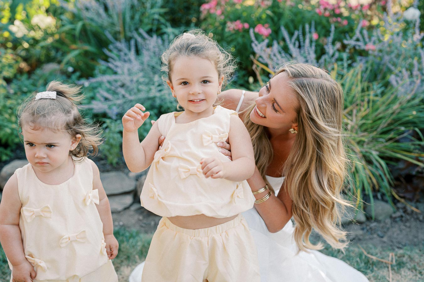A woman smiling at two young girls in matching cream-colored dresses with bows, standing in a garden filled with colorful flowers and greenery.