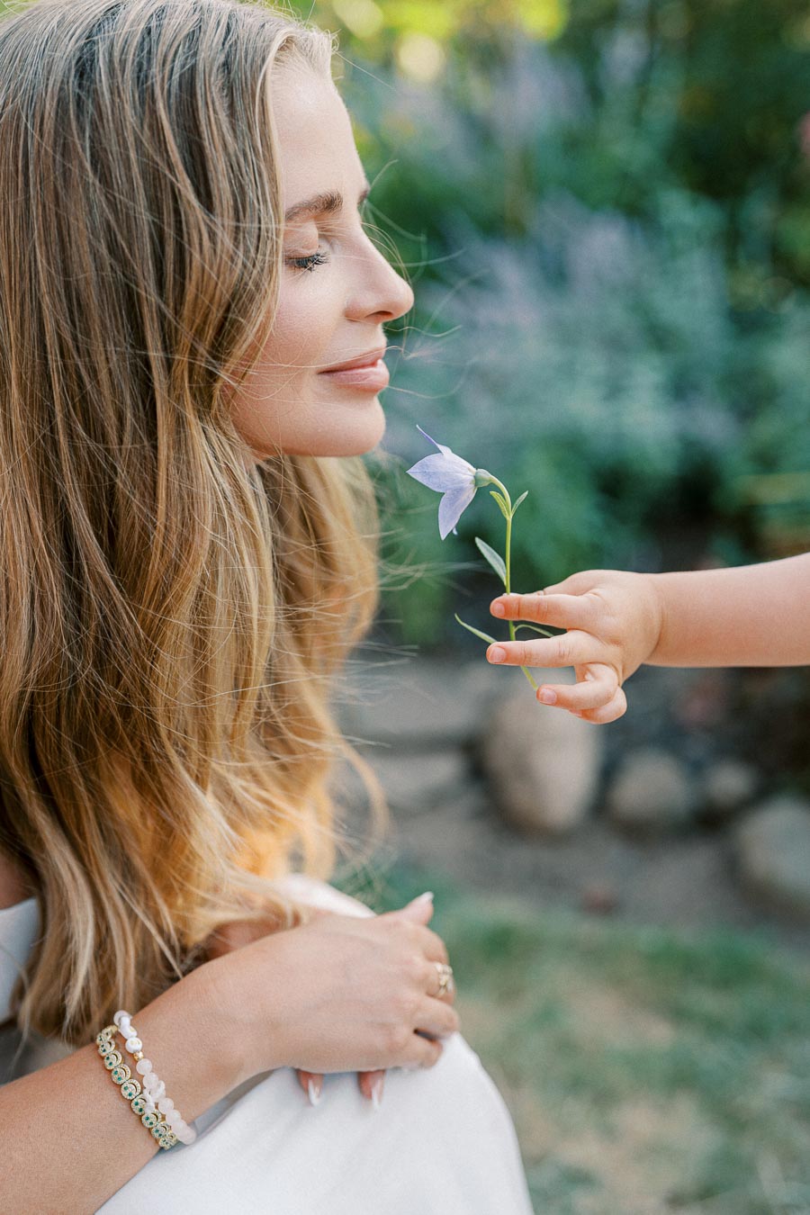 A serene woman with long blonde hair enjoys the scent of a purple flower held by a child's hand, set in a lush garden background.