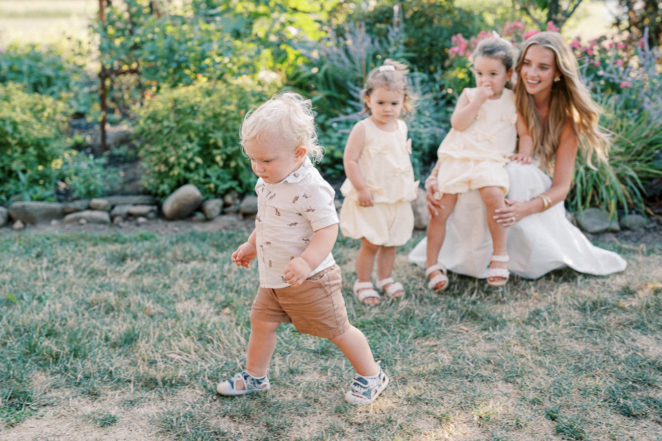 A young child walking on grass in a garden while two toddlers and a woman in a white dress sit in the background surrounded by greenery and flowers.