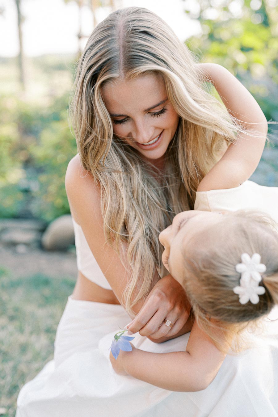 A woman with long blonde hair smiling at a child holding a blue flower, outdoors on a sunny day.