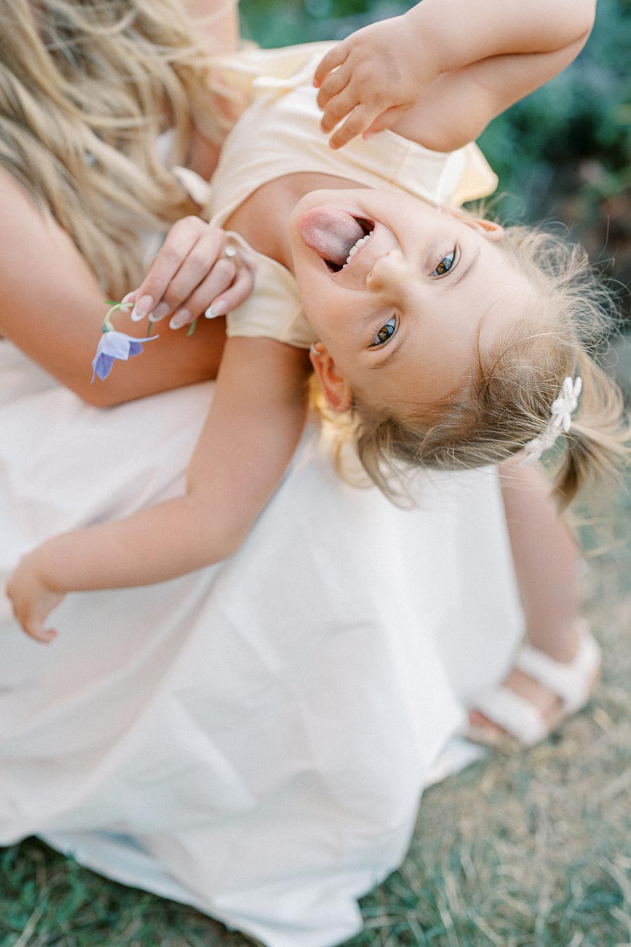 A joyful child with a playful expression, being held upside down by a woman with long blonde hair, holding a small blue flower. The scene is outdoors with a blurred green background, capturing a moment of happiness and connection.