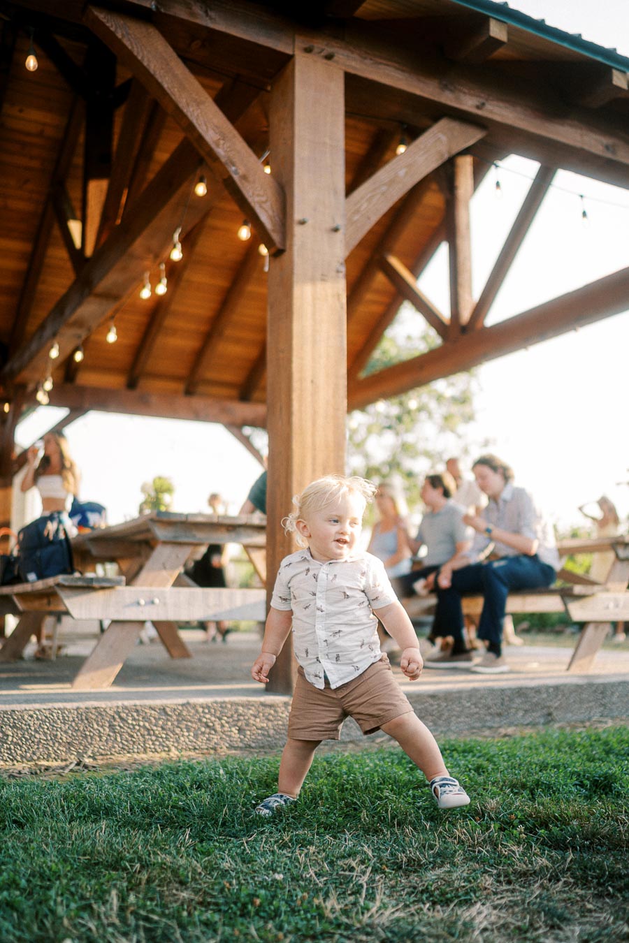 A toddler playing on grass under a wooden pavilion with people sitting at picnic tables in the background, enjoying a sunny day outdoors.