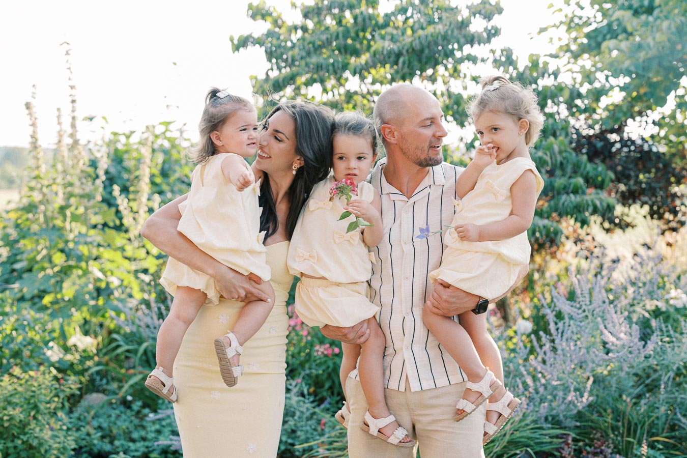 A happy family with three young children in matching yellow dresses enjoying a sunny day in a lush garden. The mother and father hold their children close, surrounded by vibrant greenery and flowers, capturing a joyful and serene moment outdoors.