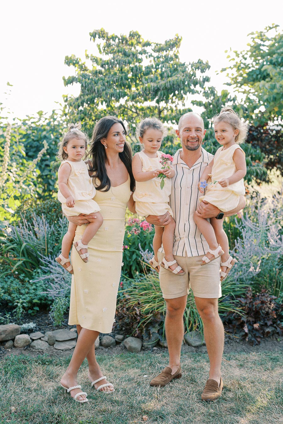 A happy family of five enjoys a sunny day in a lush garden, with parents holding their three young children, all dressed in coordinated light yellow outfits.