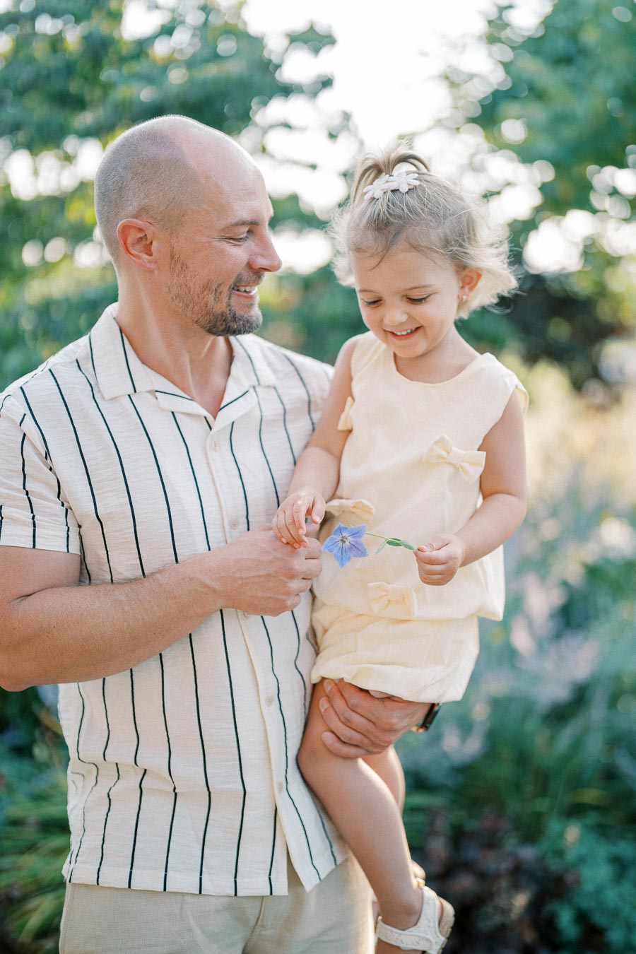 A father holding his smiling young daughter outdoors, both dressed in light summer clothing, surrounded by greenery and sunshine, emphasizing family bonding and joy.
