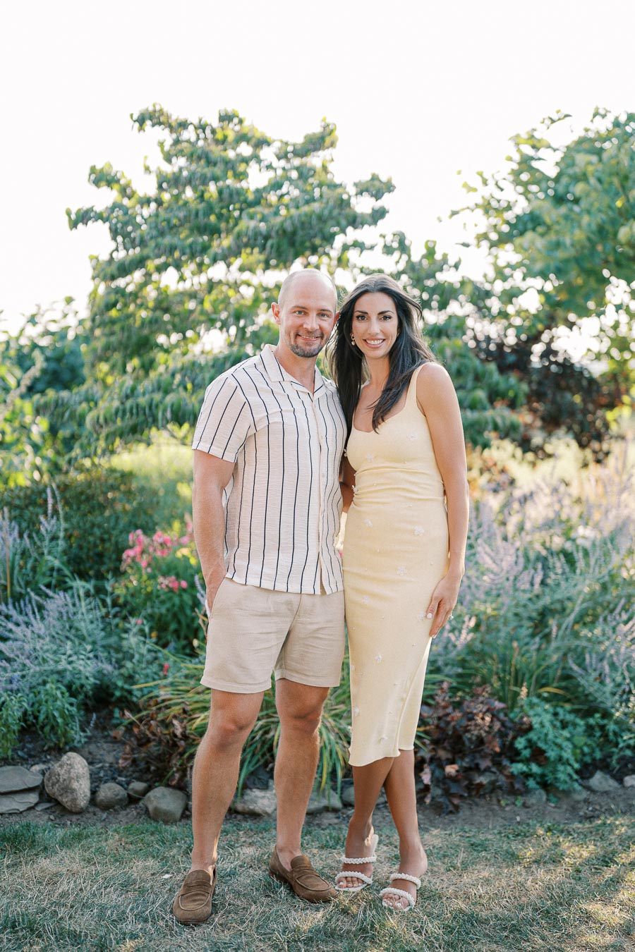 A couple smiling outdoors in a garden setting, surrounded by vibrant green foliage and colorful flowers. The man is wearing a striped shirt and beige shorts, while the woman is in a light yellow dress and sandals.