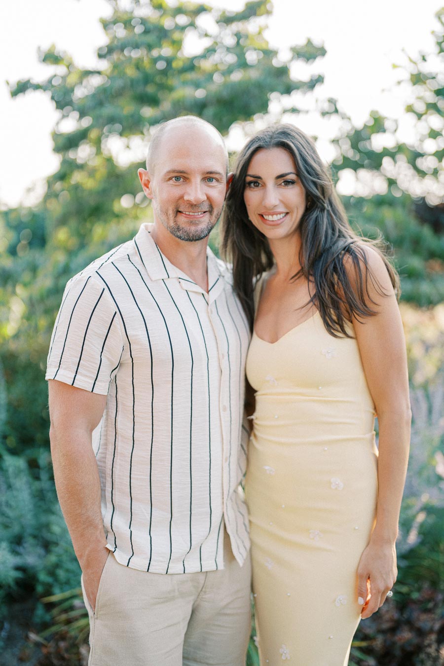 Couple posing together outdoors with green foliage in the background. The man is wearing a striped shirt, and the woman is in a yellow dress, both smiling warmly.