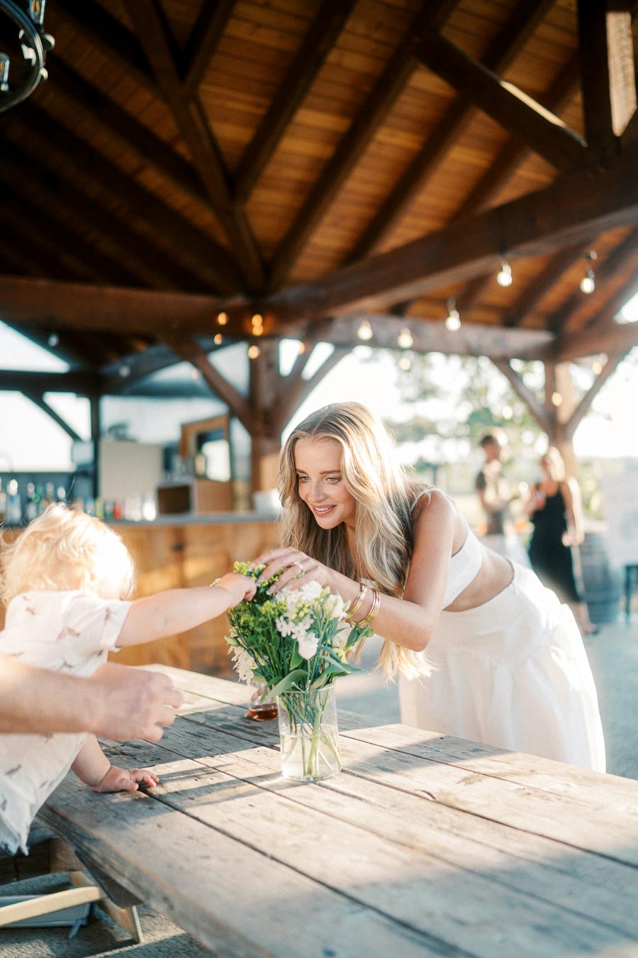 A woman in a white dress is smiling and reaching for a bouquet of flowers on an outdoor wooden table, under a canopy with string lights. A small child reaches towards the flowers. The setting suggests a warm, sunny day and an intimate gathering.