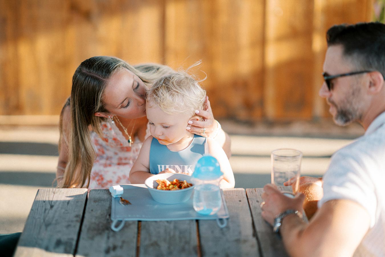 A mother affectionately kisses her young child on the head while they sit outside at a wooden table, accompanied by a man holding a glass. The child is wearing a bib labeled HUNK and is focused on a bowl of food, with a blue sippy cup nearby. The scene captures a warm family moment in natural light.