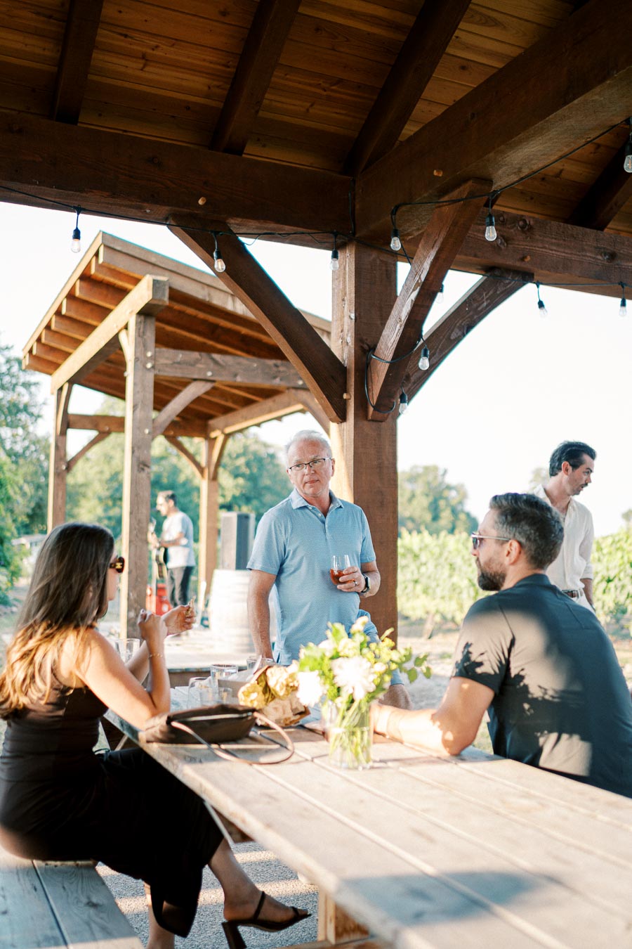 A group of people enjoying an outdoor gathering at a wooden picnic table under a rustic pavilion, with sunlight filtering through the trees in the background.