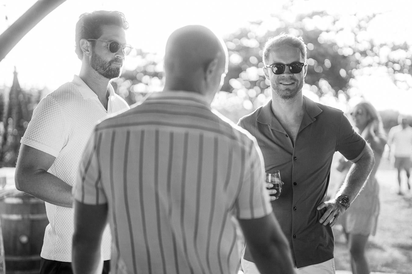Black and white image of three men in sunglasses at an outdoor event. They are casually dressed and engaged in conversation under bright sunlight, with blurred natural scenery in the background. The man on the right is holding a drink, suggesting a social gathering or celebration.