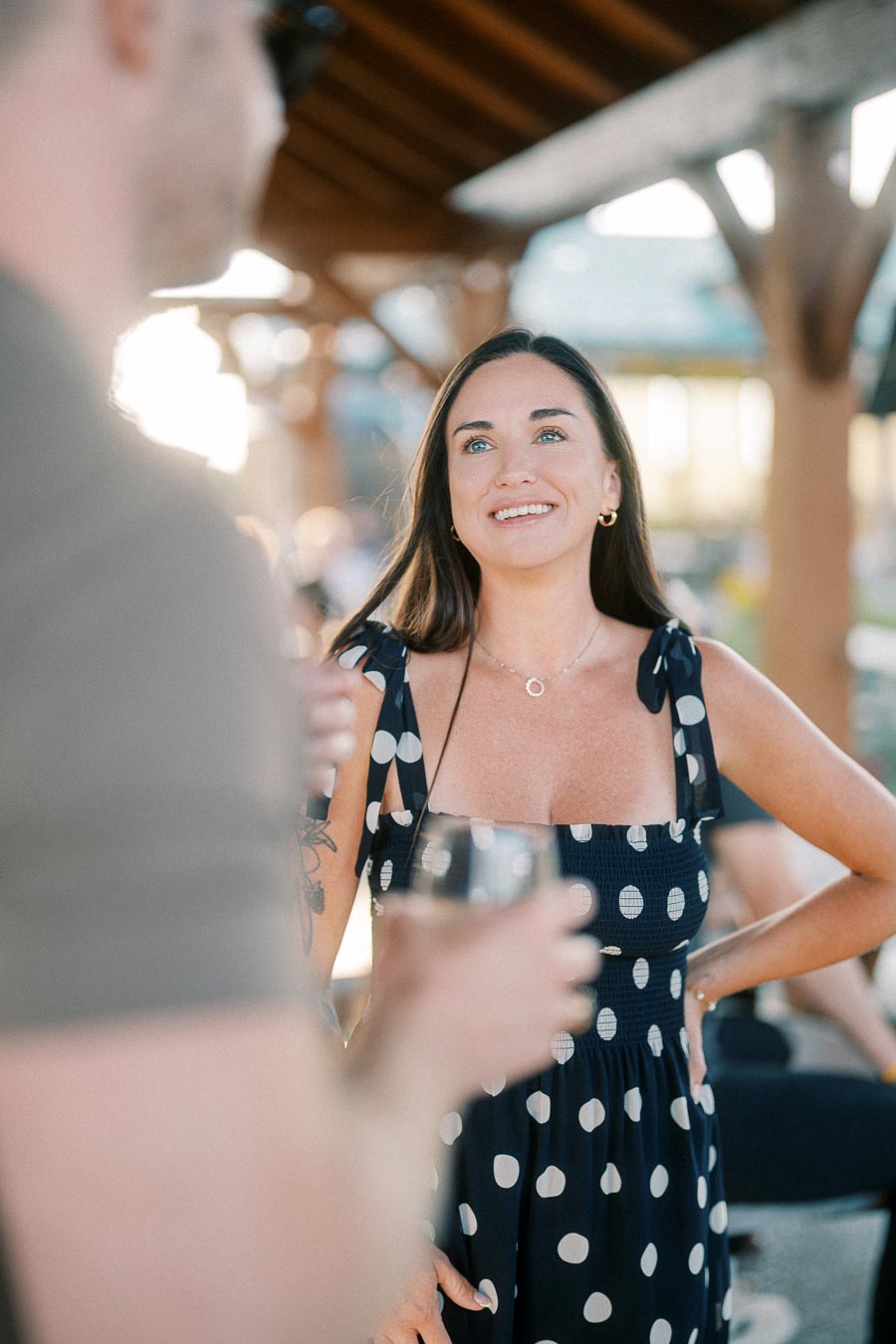 A woman in a polka dot dress smiles and engages in a conversation at an outdoor social event, holding a wine glass in natural sunlight.