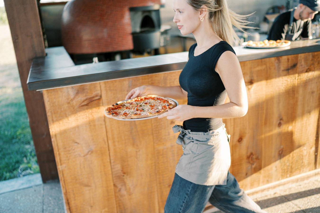 A woman with a ponytail carrying a freshly baked pizza past a wood-fired oven in an outdoor pizza restaurant.