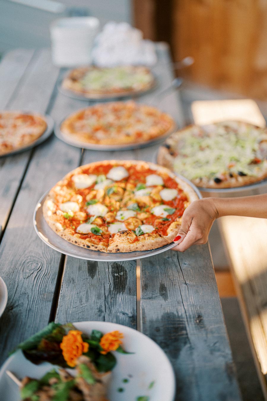 Close-up of a freshly baked pizza topped with mozzarella cheese and fresh basil being placed on a rustic wooden table, surrounded by other assorted pizzas and a salad, capturing an outdoor dining experience.