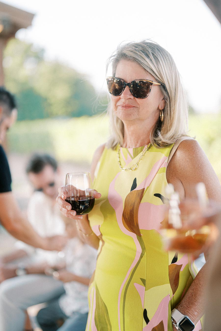 Woman in a vibrant floral dress and sunglasses enjoying a glass of wine outdoors.