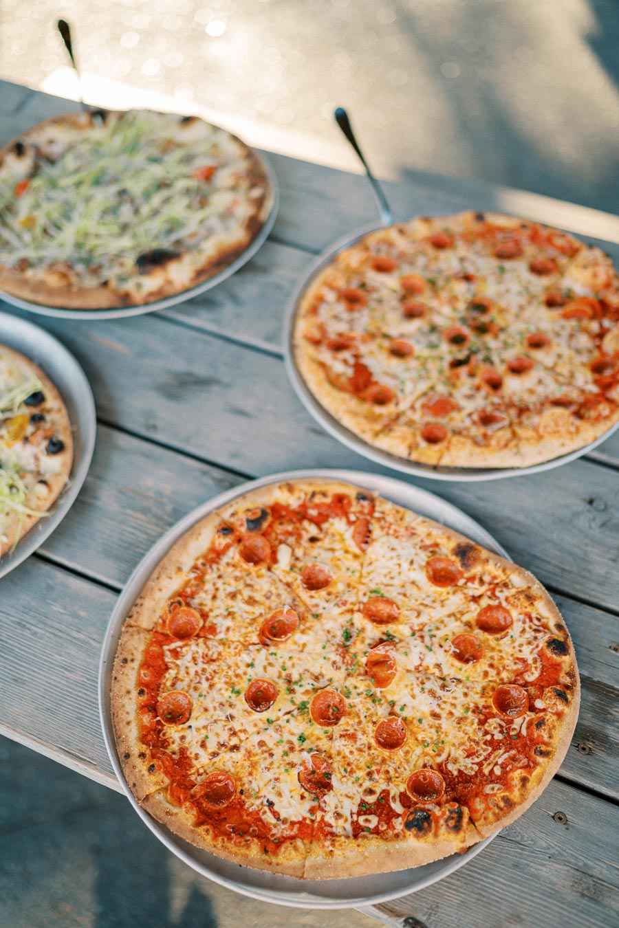 Four delicious pizzas with various toppings on metal trays placed on a rustic wooden table. Perfect for outdoor dining or sharing with friends and family.
