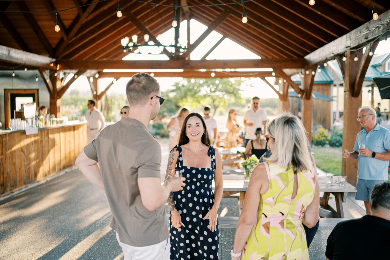 Outdoor social gathering under a wooden pavilion, with people mingling and enjoying a sunny afternoon. The image captures the relaxed atmosphere of a summer event, featuring individuals in casual attire, surrounded by natural scenery and festive lighting.