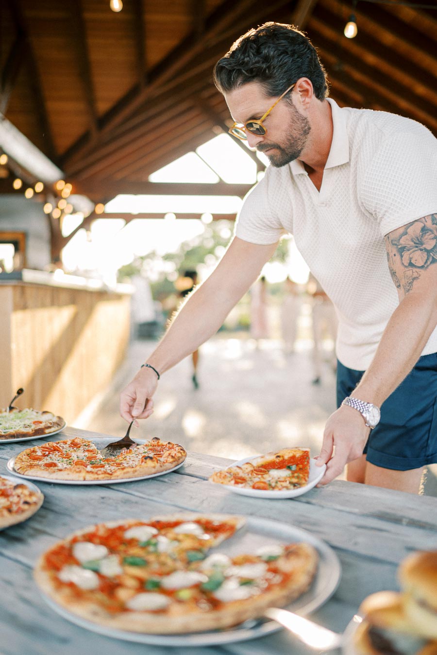 A man selecting a slice of pizza from a table spread with various pizzas in an open, rustic setting under wooden beams, wearing casual summer attire and sunglasses, suggesting a relaxed outdoor gathering or party.