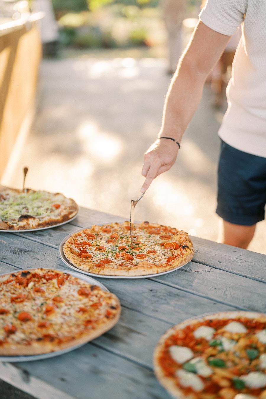 A person slicing a freshly baked pepperoni pizza with a fork on a rustic wooden picnic table, surrounded by various other pizzas, in a sunny outdoor setting.