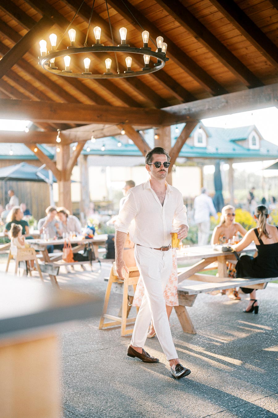 Outdoor social gathering under a wooden pavilion, featuring a stylish man in a white outfit and sunglasses, walking with a drink in hand. He is surrounded by people seated at wooden picnic tables, enjoying a sunny day. Above, a modern chandelier adds to the relaxed, elegant atmosphere.