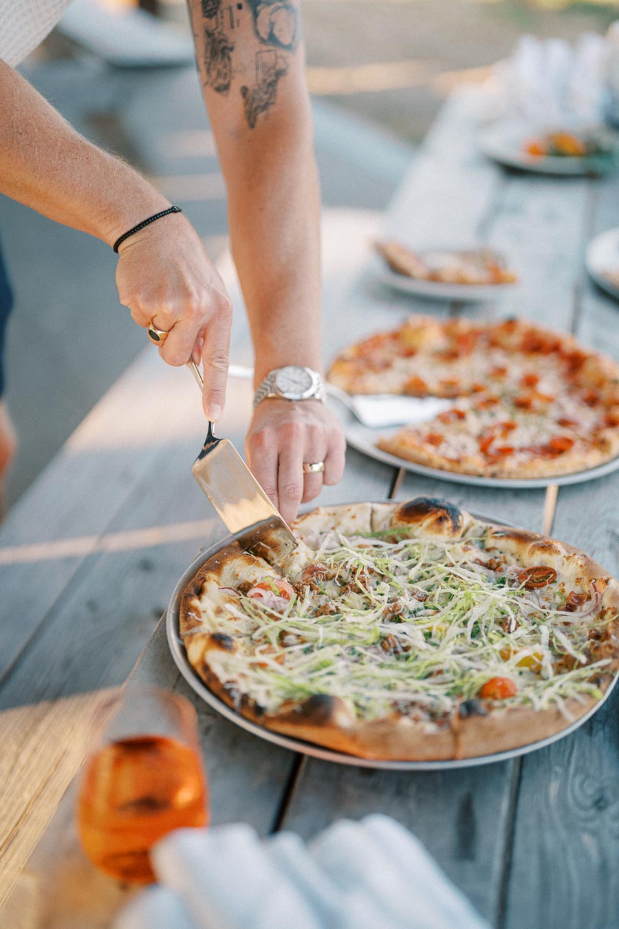 A person slicing a freshly baked pizza with a spatula on a wooden picnic table, featuring toppings like shaved vegetables and tomatoes. Additional pizzas and a glass of iced tea are visible in the blurred background, suggesting an outdoor gathering or picnic setting.