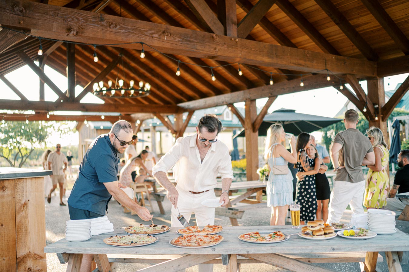 Group of people enjoying an outdoor gathering under a wooden pavilion, with two men serving pizza onto plates from a table laden with food, including burgers, appetizers, and drinks. Others socialize in the background, creating a lively and festive atmosphere.