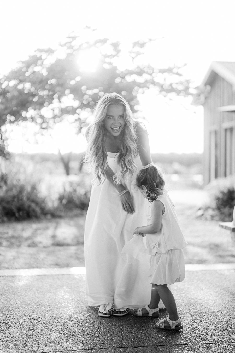 A joyful woman in a flowing white dress plays with a young girl outdoors on a sunny day, with trees and a rustic building in the background.