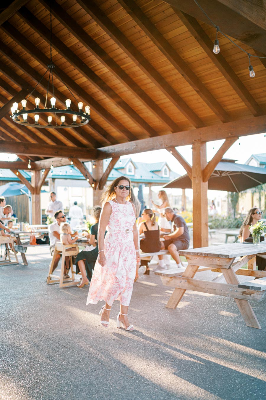 A woman in a floral dress and sunglasses walks under a wooden pavilion at an outdoor gathering, with seated people socializing at picnic tables in the background.