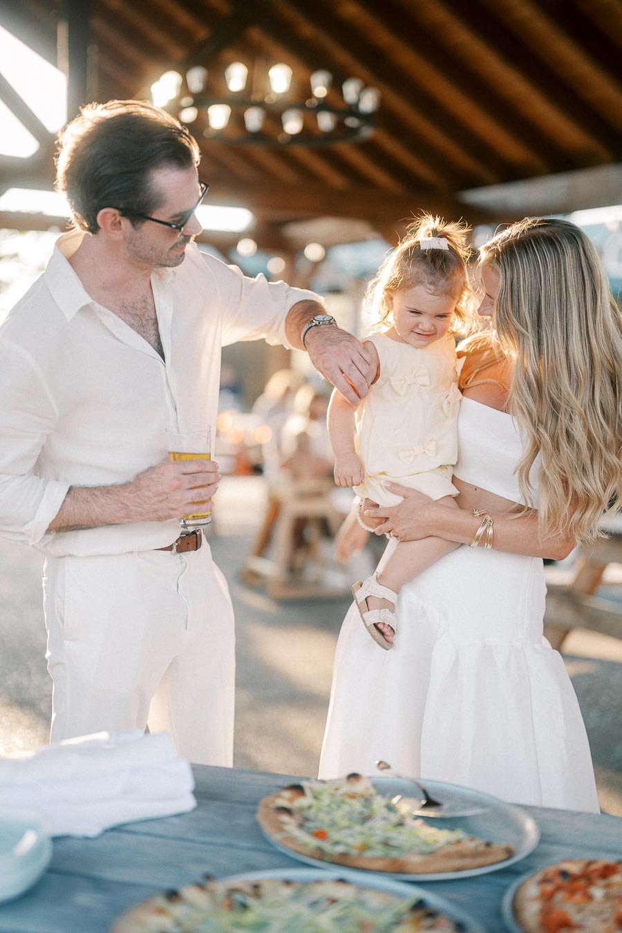 A family enjoying a sunny outdoor gathering, with a father holding a drink, a mother holding a smiling toddler, and pizzas on a table in the foreground.