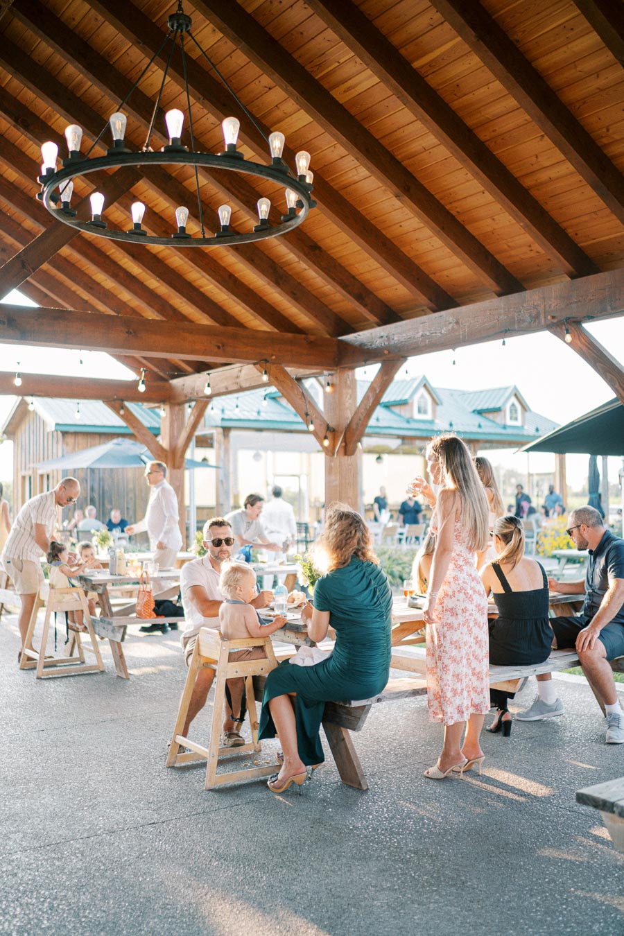 Outdoor gathering under a wooden pavilion with a chandelier, featuring people enjoying a meal and conversations at wooden tables, surrounded by a sunny and relaxed atmosphere.
